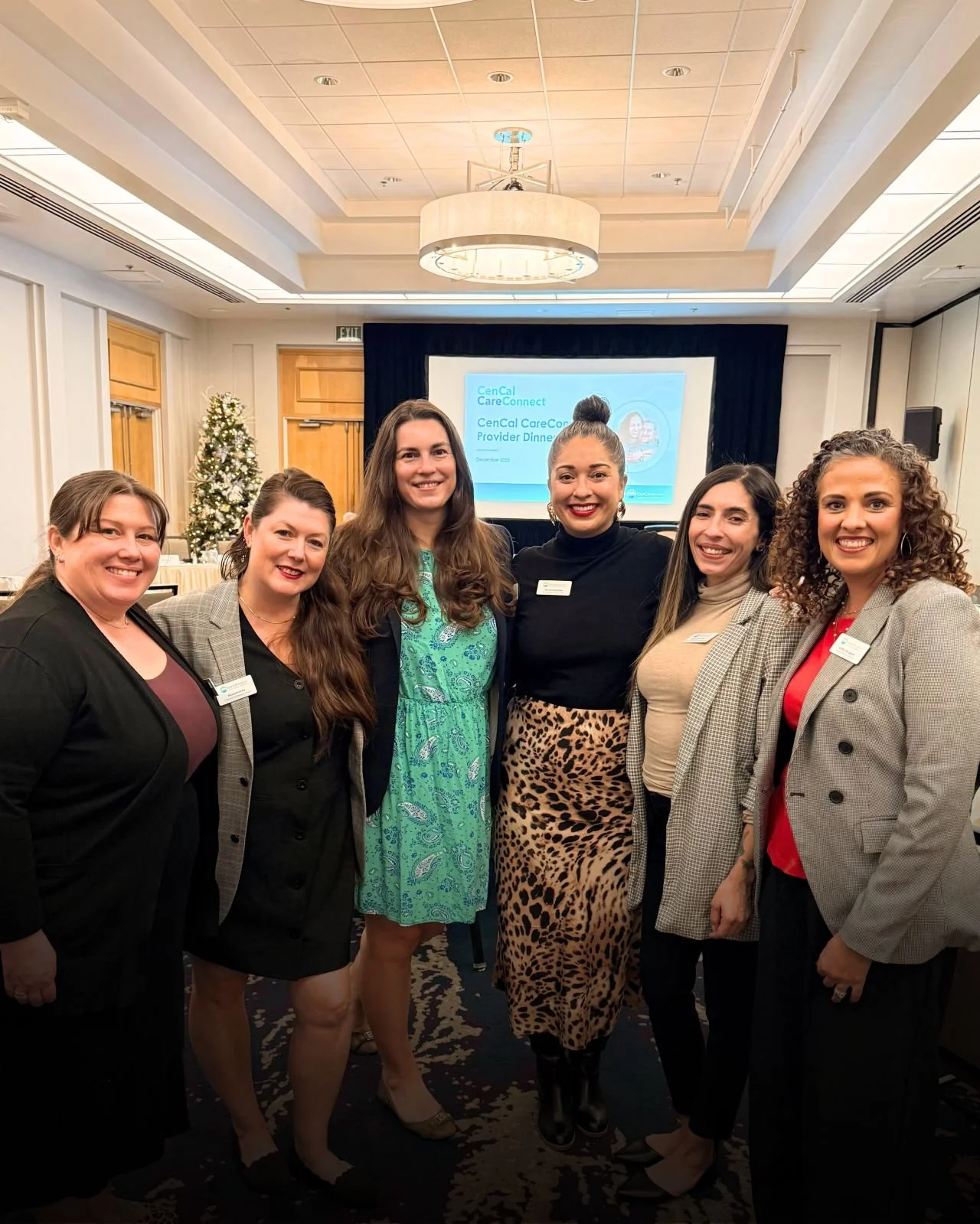 Group of six women standing together in a conference room, smiling at the camera, with a Christmas tree and a presentation screen in the background.