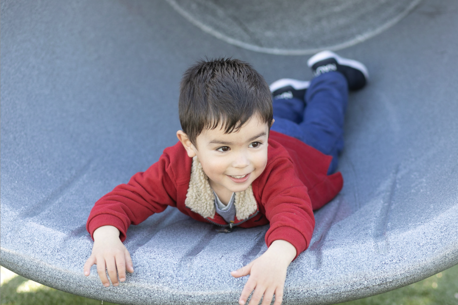 A young boy lying on his stomach on a playground slide, smiling and looking to the side.