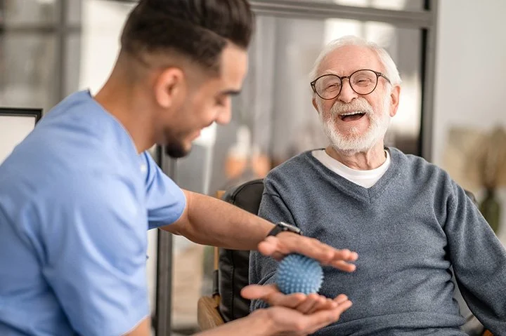 A healthcare worker in blue scrubs feeding a senior man a blue textured ball.