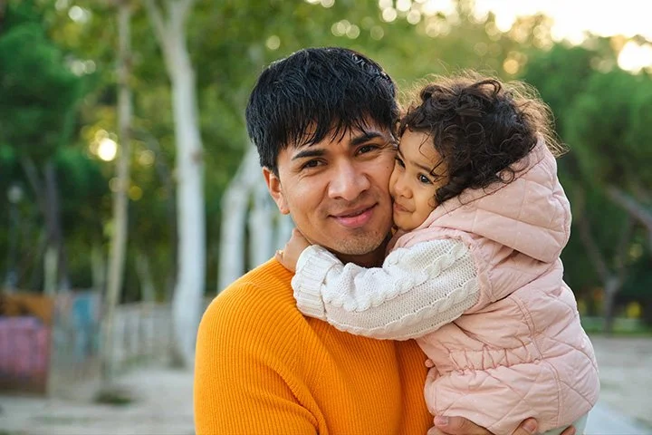 A man holding a young girl, both smiling and embracing outdoors in a park with trees in the background.