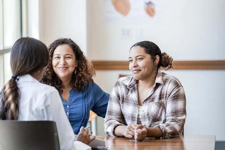Three women sitting at a table engaged in conversation, with a light-colored wall in the background.