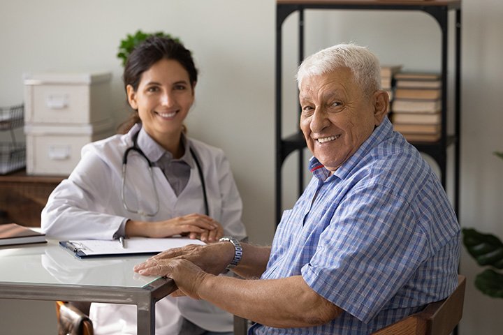 A smiling female doctor wearing a white coat and stethoscope along with an older male patient sitting at a desk in a medical office.