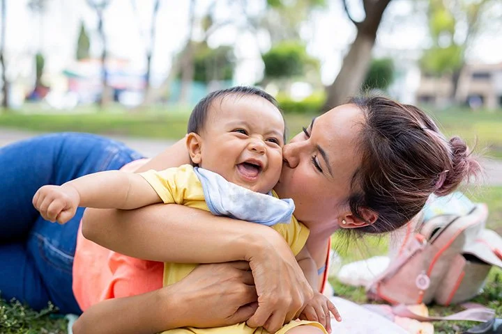 A woman laying on the grass outside, hugging and kissing a smiling baby who is laughing and reaching out.