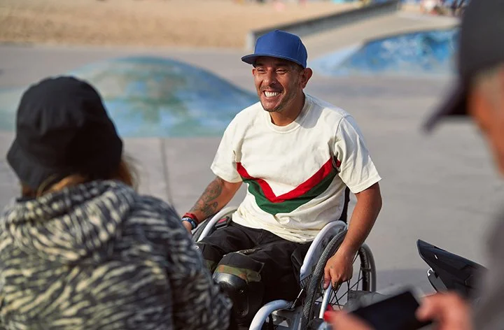 A man in a wheelchair smiling and talking to two people outdoors near a skate park.