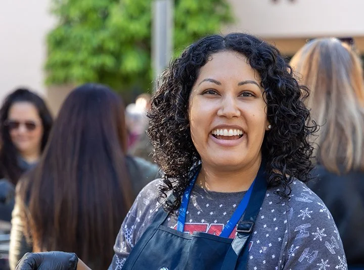 Smiling woman in a crowd, wearing a dark apron and a Christmas-themed shirt, outdoors.