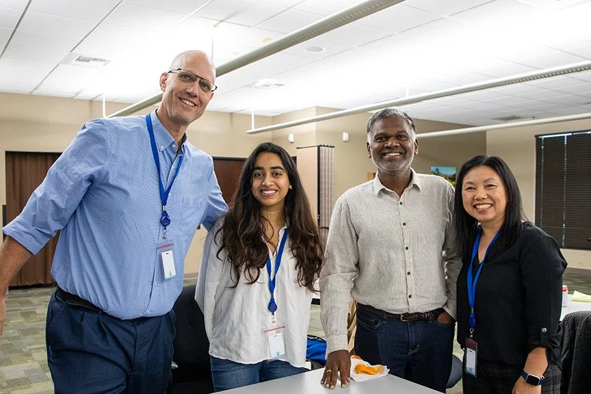 Four diverse professionals standing together in an office or conference room, smiling for the camera.