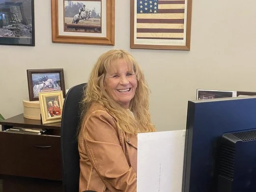 A woman with long blonde hair and a tan jacket sitting at a desk, smiling, with framed pictures and a picture of the American flag on the wall behind her.