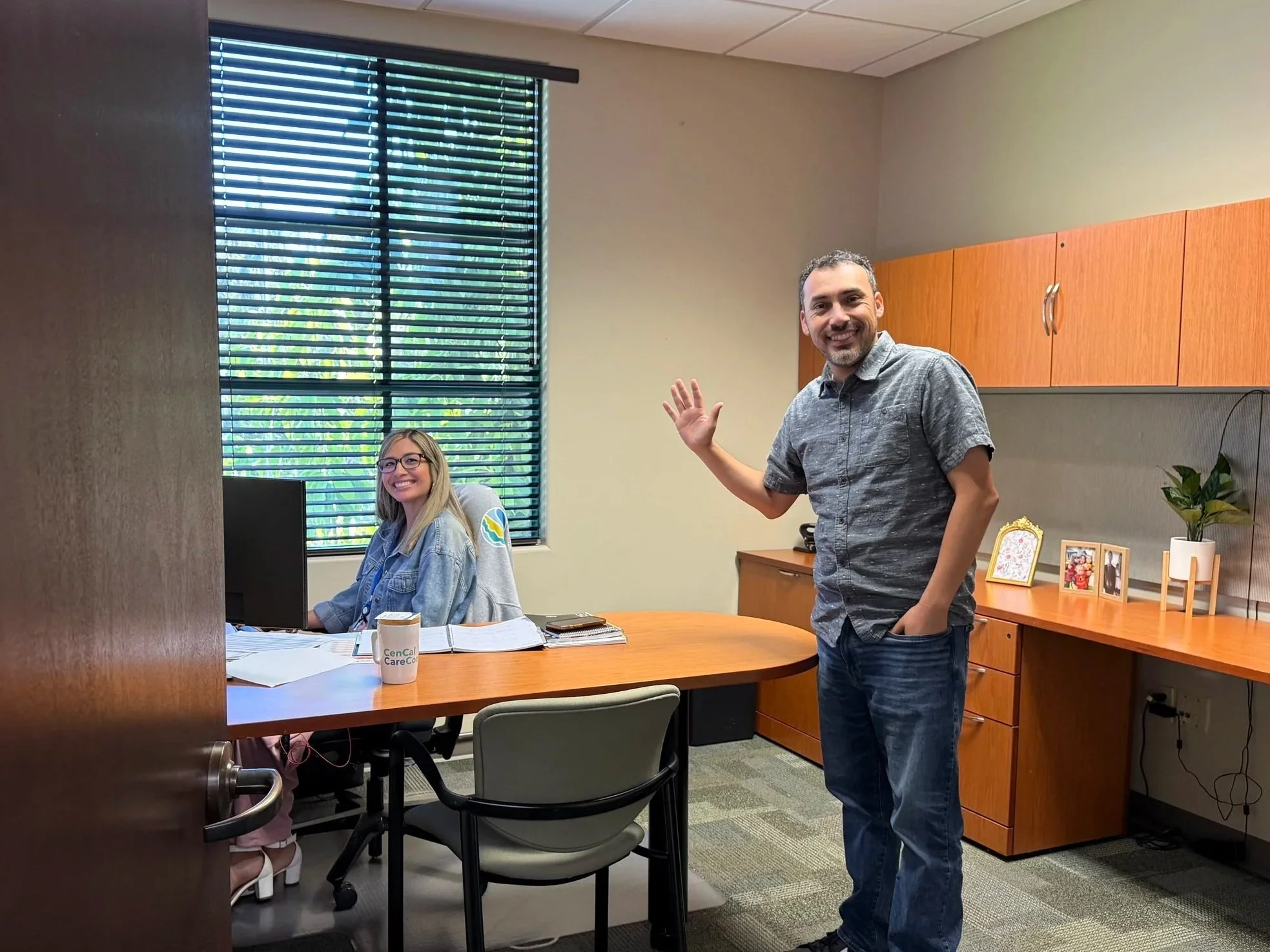 Two smiling people in an office, one woman sitting at a desk with a computer and the other man standing and waving