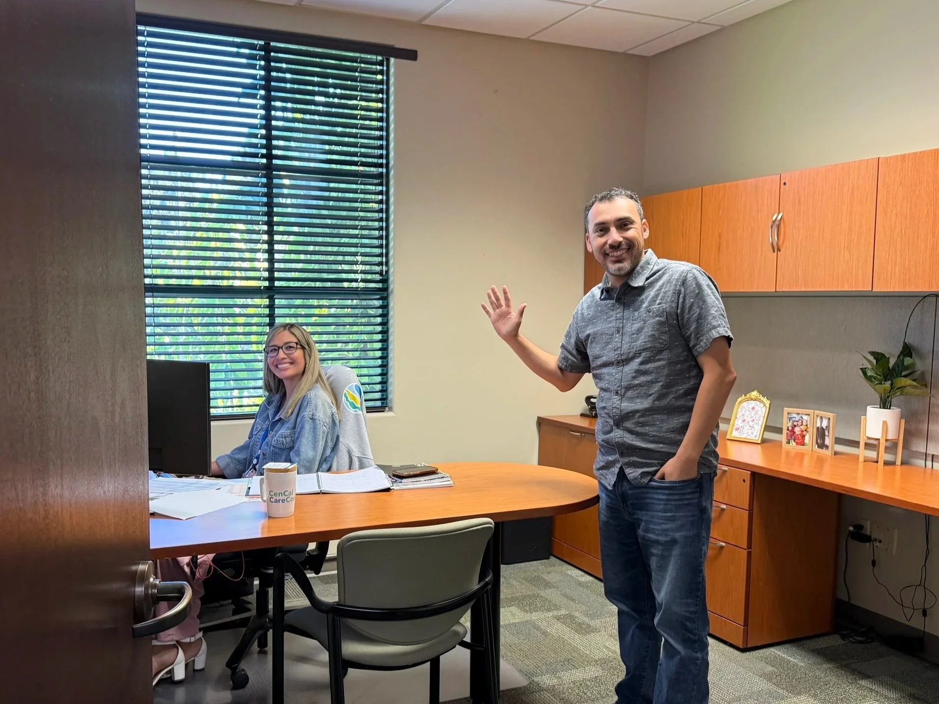 A woman with glasses and a man smiling in an office, woman seated at desk and man standing, both looking at the camera. The office has a window, desk, and decorative photos.