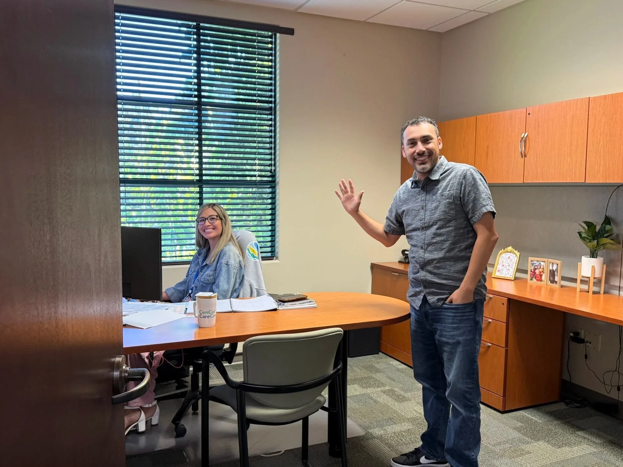 A man waves and smiles in an office room where a woman sits at a desk with a computer, papers, and a coffee mug. The room has a window with blinds and wooden cabinets.