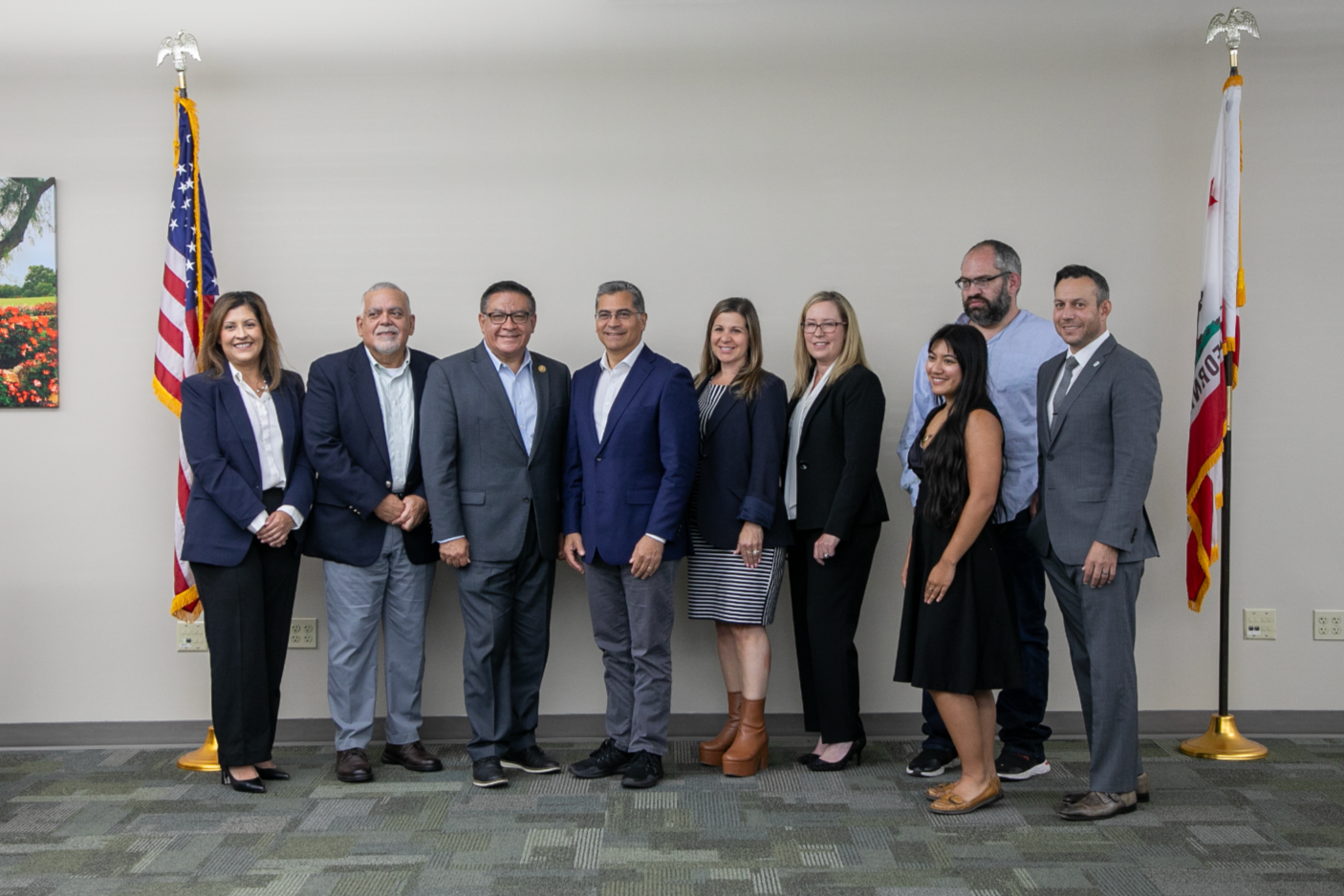 A group of nine diverse professionals posing together in a conference room with American and California flags behind them.