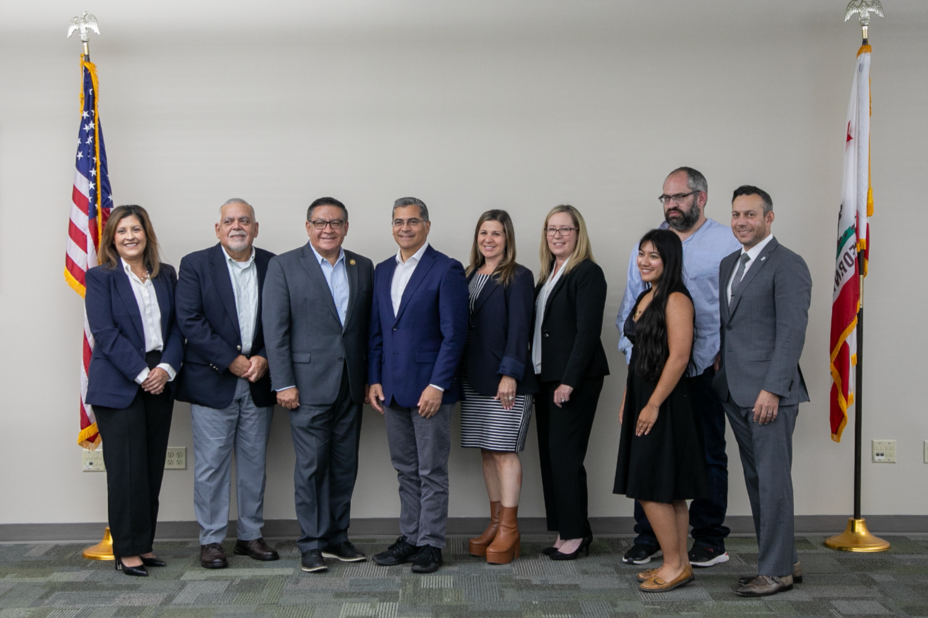 A group of ten professionally dressed people standing in a line inside a room with two American flags on poles, on either side of the group.