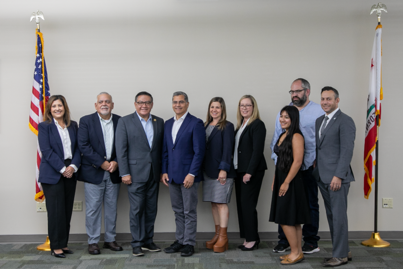 A group of nine diverse professionals standing in a row in front of two American flags and a plain wall, smiling at the camera.