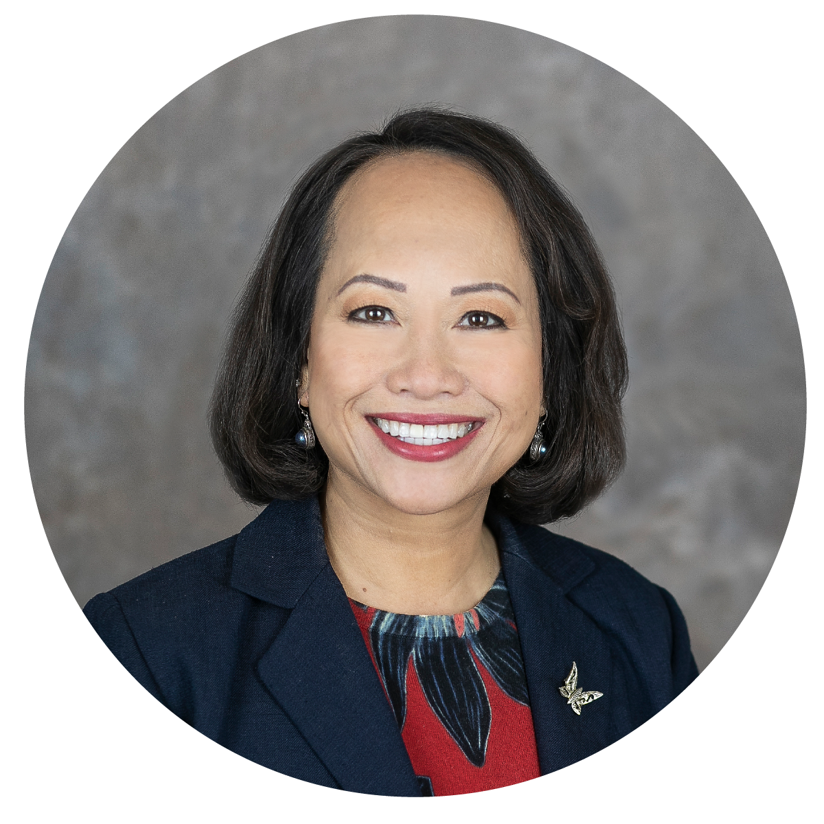 Professional headshot of a woman with shoulder-length dark hair, wearing a dark blazer, a colorful top, and a butterfly pin, smiling against a neutral background.