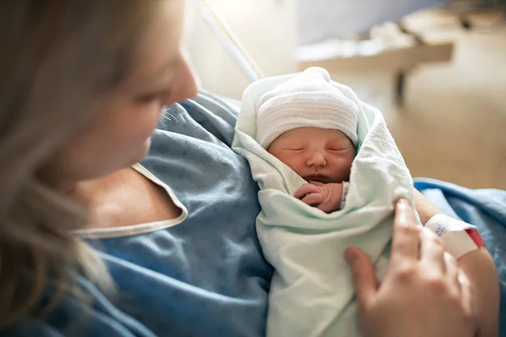 Newborn baby wrapped in a white blanket and wearing a striped cap, being held close by a woman in a hospital gown.