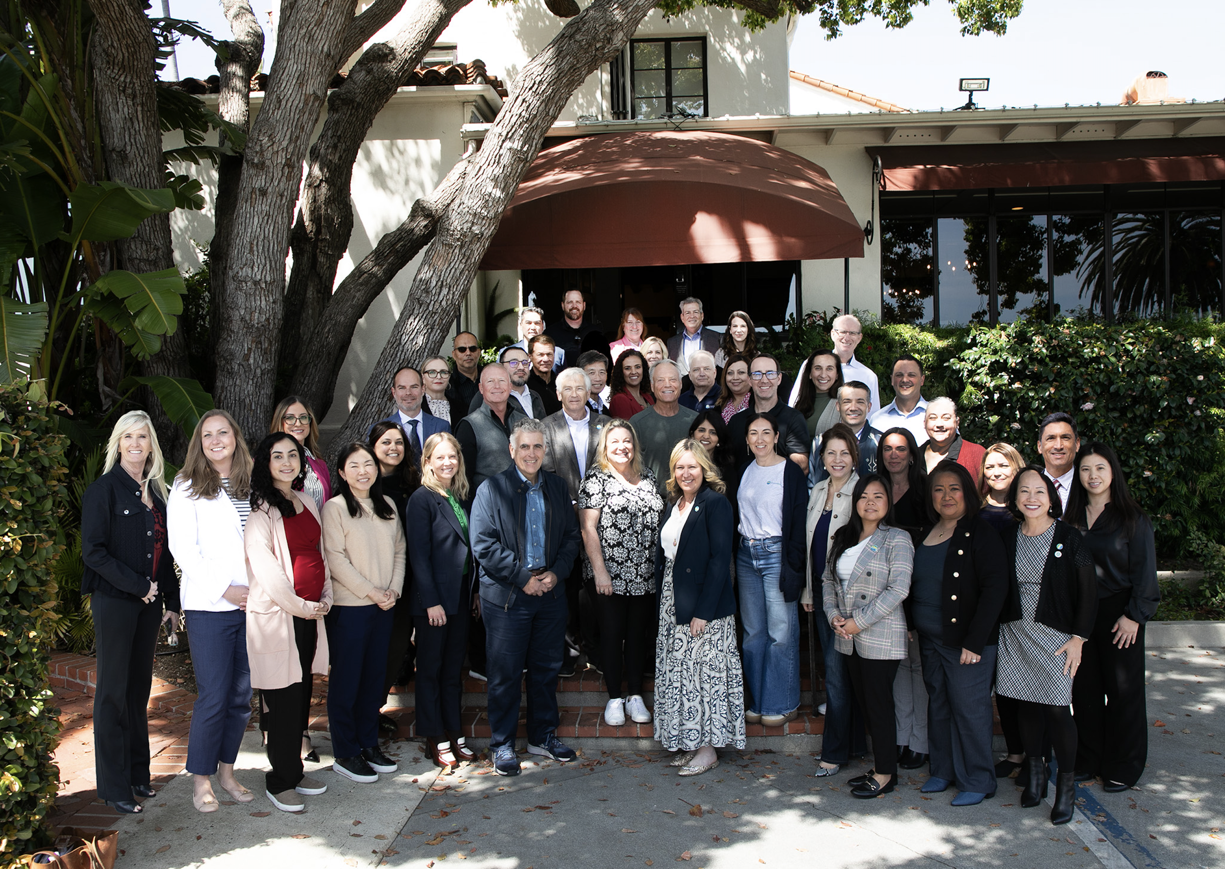Group photo of approximately 40 diverse people standing on steps outside of a building with greenery and trees around.