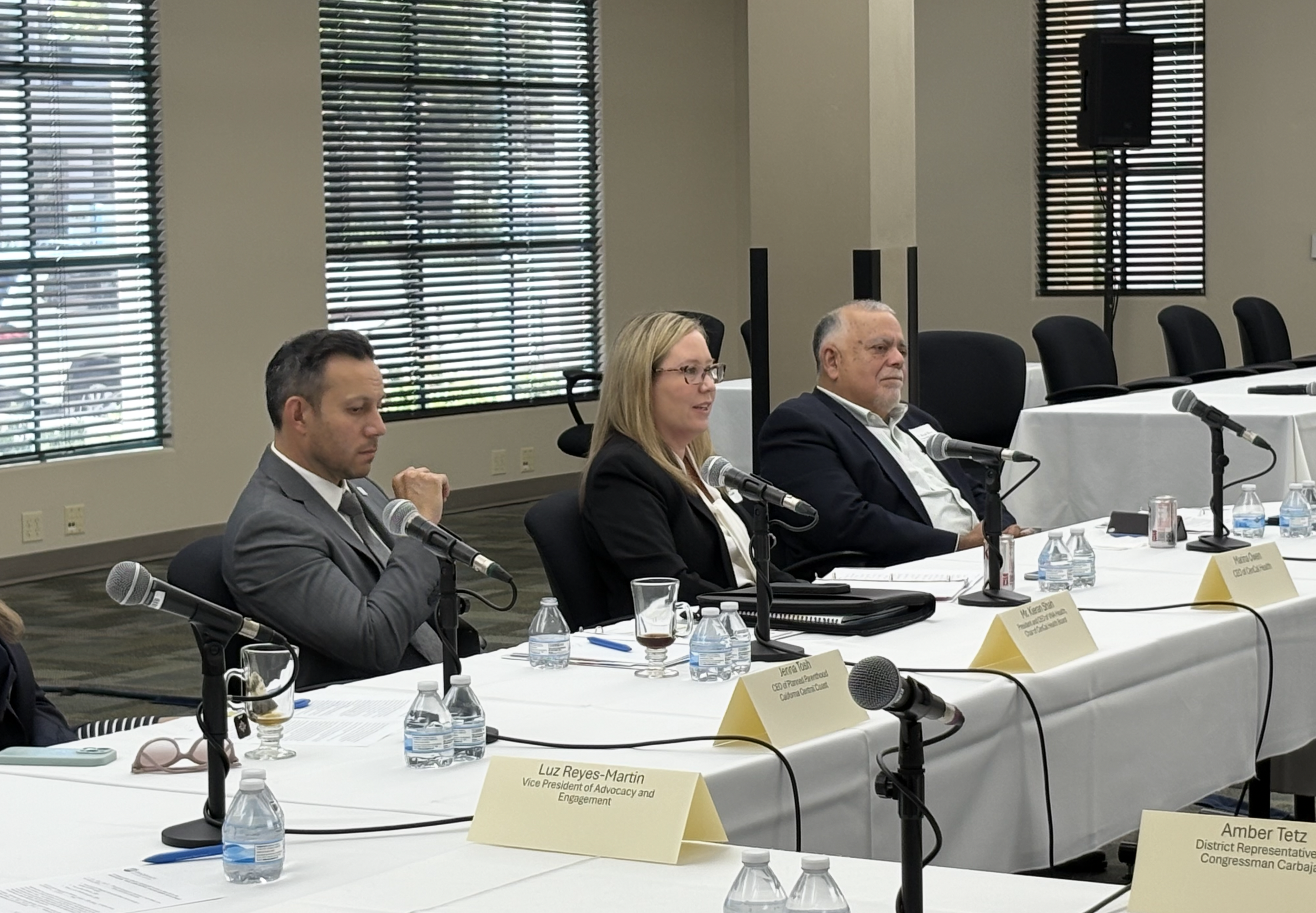 Three people sitting at a long table with microphones, water bottles, and name tags in a conference room with windows and blinds.