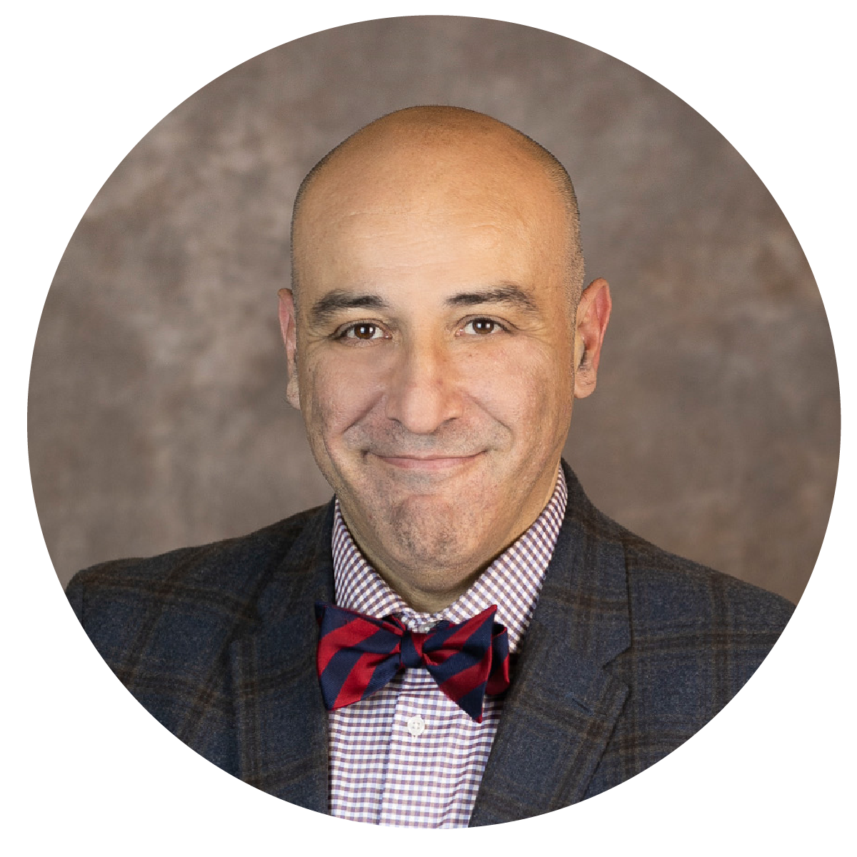 Professional portrait of a bald man smiling, wearing a checkered shirt, a plaid blazer, and a red and navy bow tie, against a gray background.