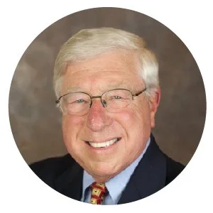 A smiling elderly man with white hair and glasses, wearing a suit, light blue shirt, and colorful tie, against a neutral background.
