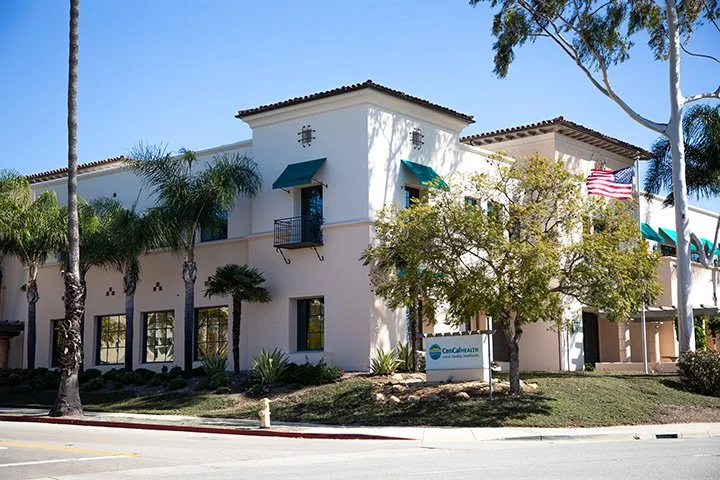 White two-story healthcare building with teal awnings, surrounded by palm trees and greenery, with a sign reading 'CenCal Health' near the sidewalk.