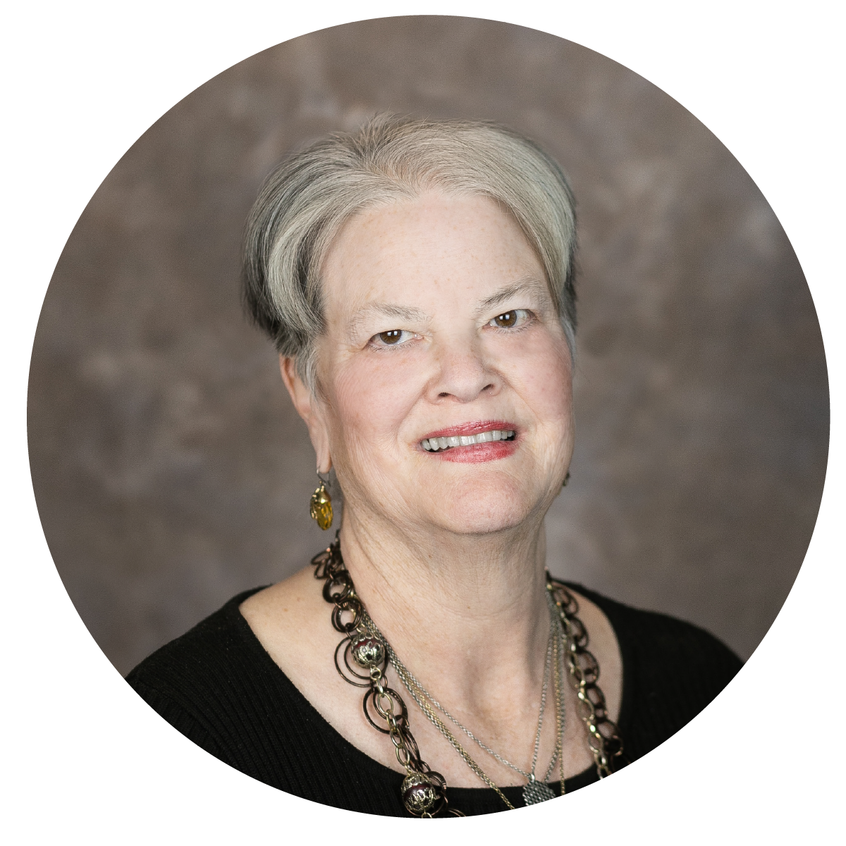 Portrait of an elderly woman with short gray hair, wearing a black top and layered necklaces, smiling at the camera against a blurred gray background.