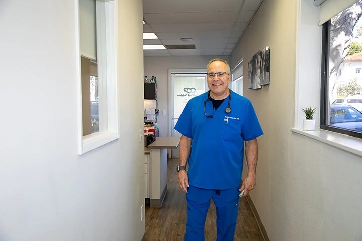 A smiling male healthcare professional in blue scrubs with a stethoscope around his neck, standing in a clinic hallway, with windows on the right and medical equipment in the background.