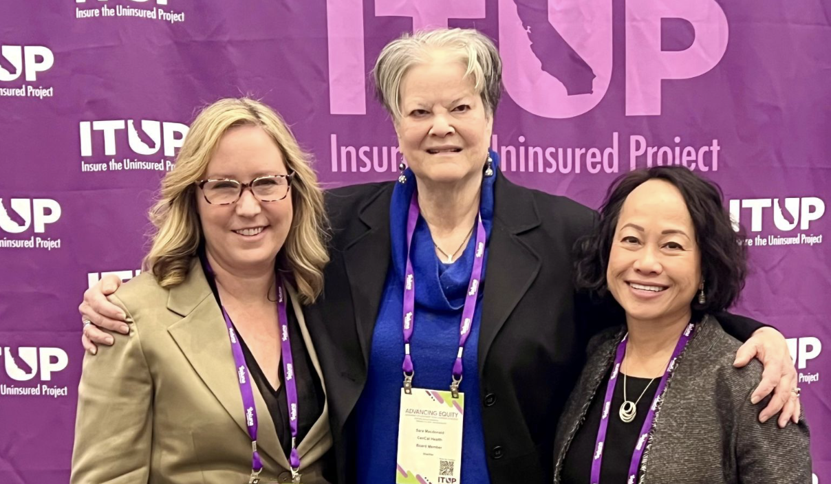 Three women smiling at a conference with a purple banner in the background that reads "ITUP Insurance the Uninsured Project." The women are wearing conference badges and lanyards.