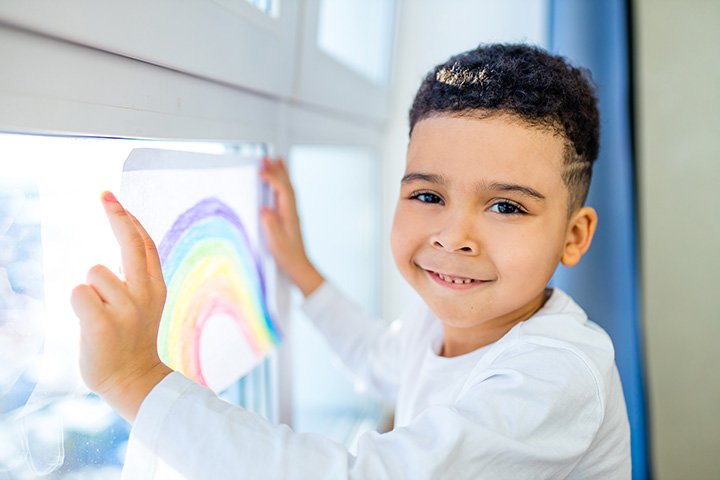 A young boy with curly hair and a white shirt smiling while pointing at a rainbow drawing on the window.