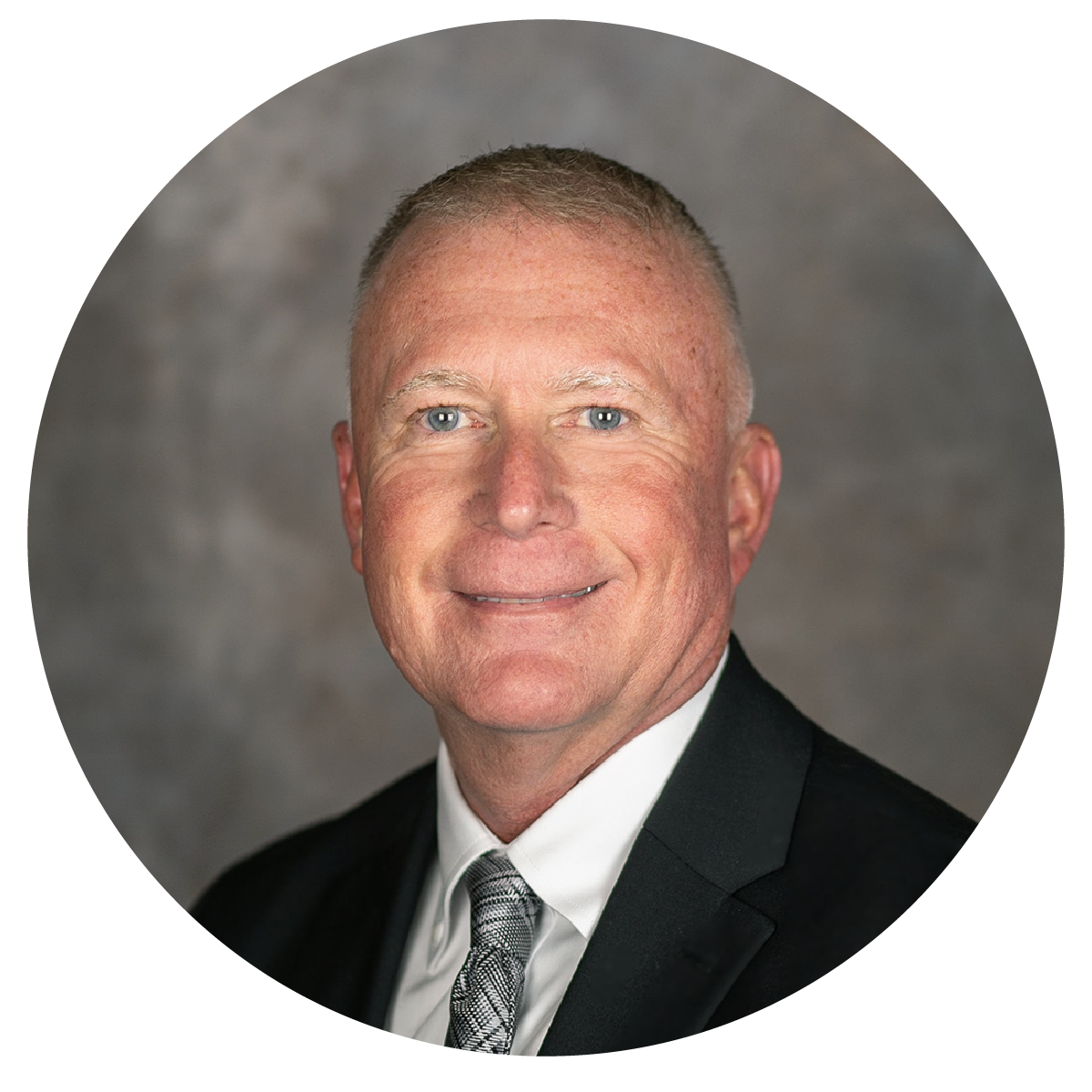 Professional headshot of a smiling middle-aged man with short light-colored hair, blue eyes, wearing a black suit, white shirt, and patterned tie, set against a neutral gray background.