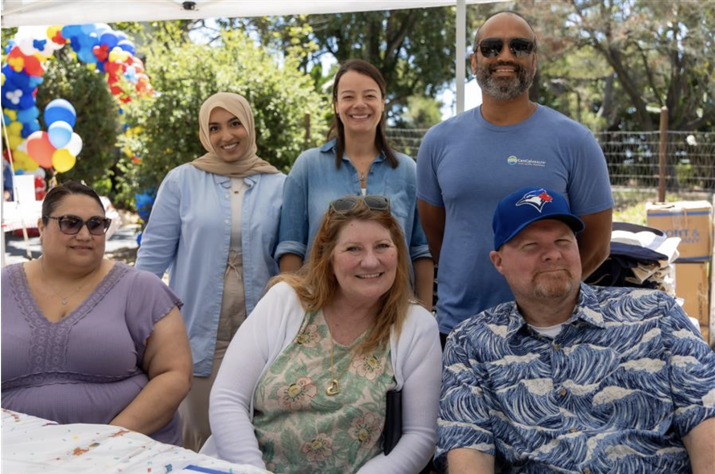 Group of six diverse adults smiling outdoors at a celebration, with colorful balloons and trees in the background.