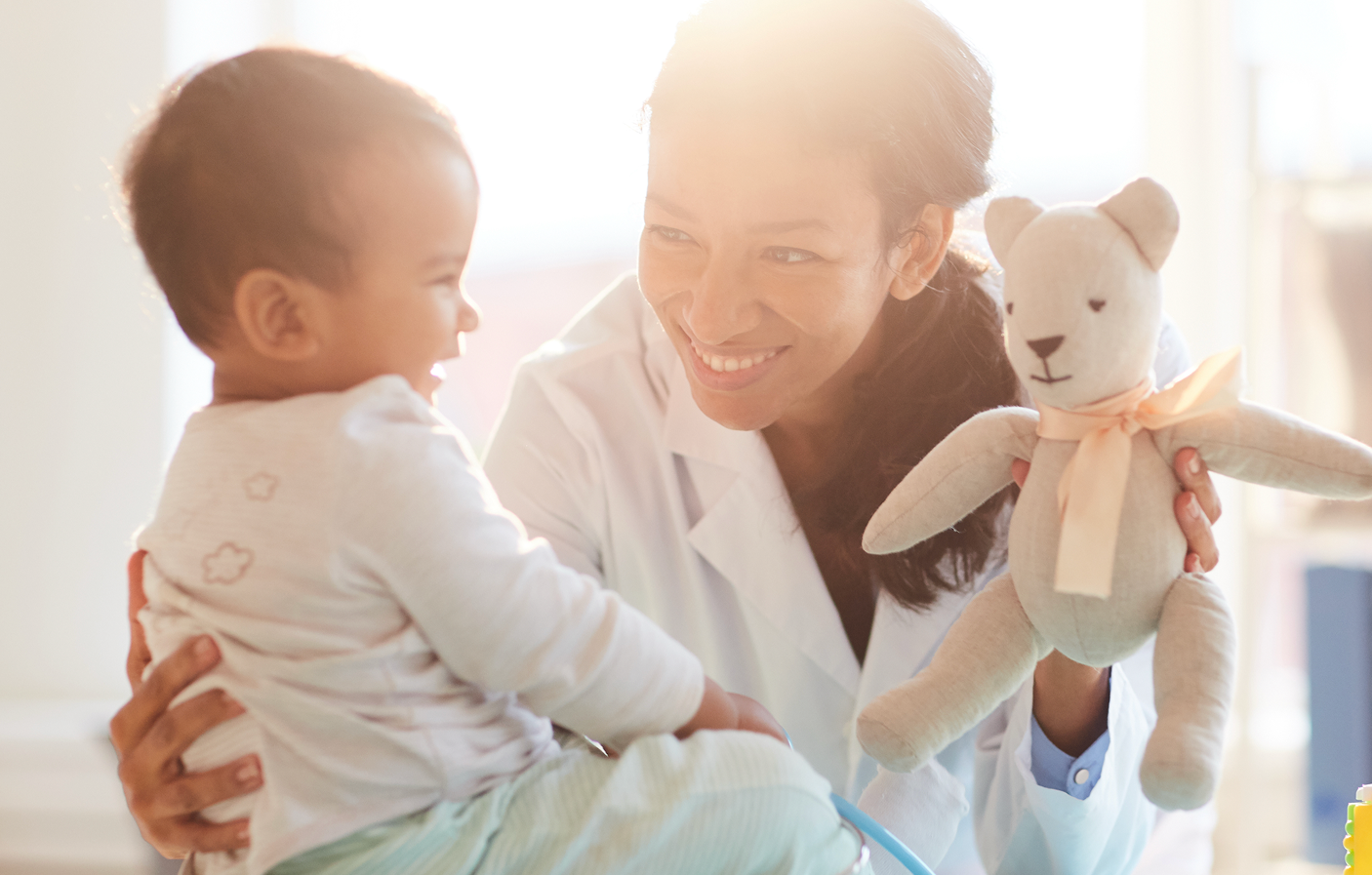 A woman, possibly a doctor, smiling and holding a teddy bear for a young child who is sitting and playing with her in a bright room.