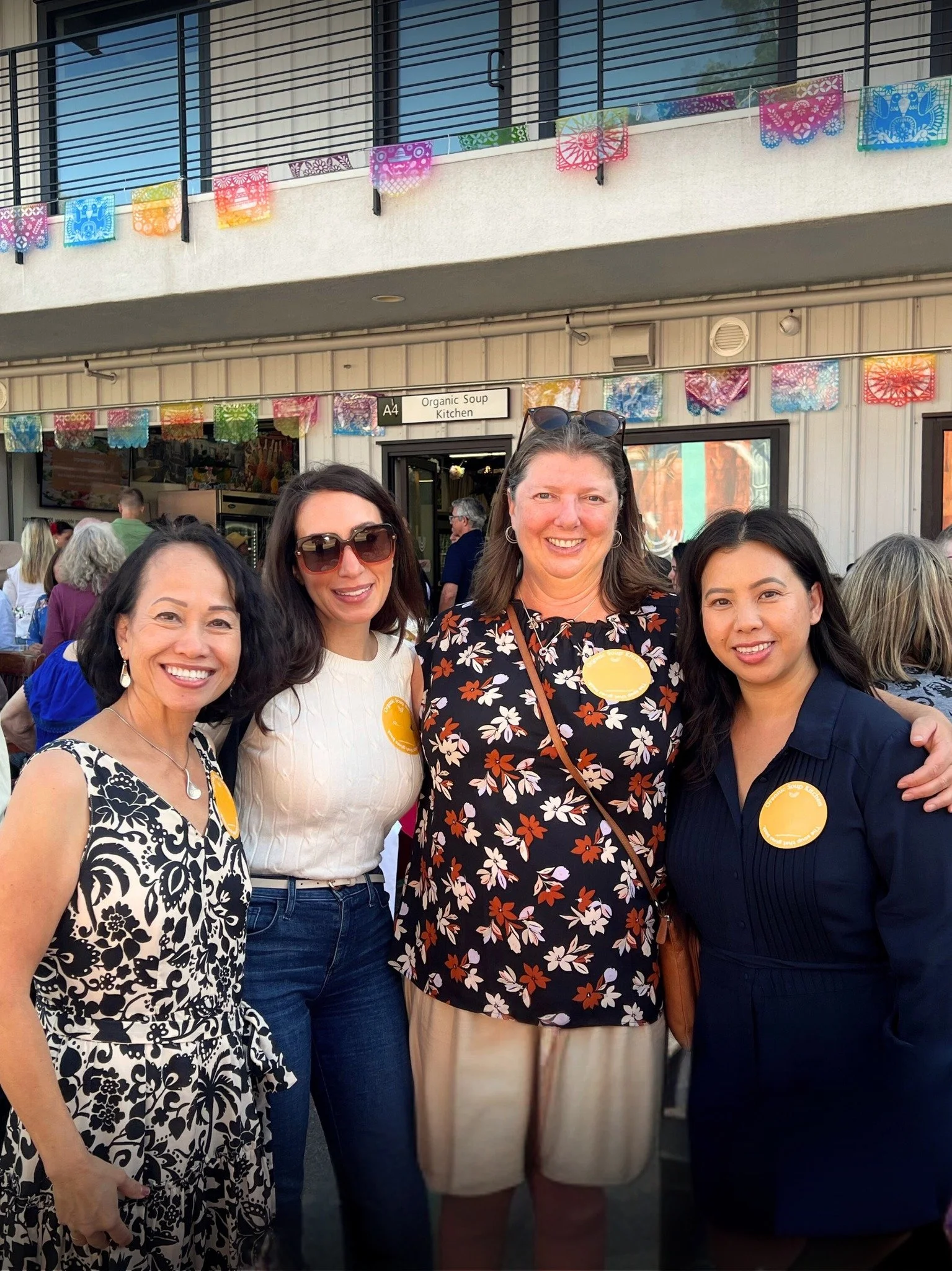 Four women standing together outdoors in front of a building with colorful papel picado decorations, smiling at the camera.