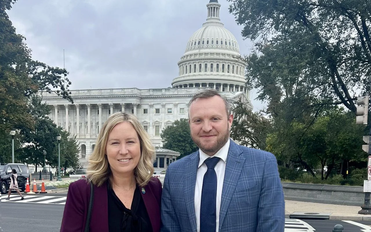 A woman and a man standing in front of the United States Capitol building in Washington, D.C. during the daytime with cloudy weather. The woman has blonde hair and is wearing a dark top with a burgundy blazer. The man has short hair and a beard, and is wearing a blue plaid suit and tie. There are trees, a street, and people in the background.