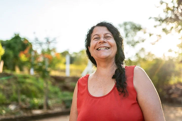 A woman with long dark curly hair is smiling outdoors, wearing a sleeveless red top, with trees and a bright sky in the background.
