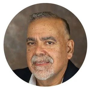 Close-up portrait of a middle-aged man with short gray hair, beard, and weathered skin, wearing a dark suit jacket and white shirt, against a gray background.