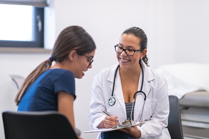 A female doctor with glasses, wearing a white coat and stethoscope, smiling while talking to a young girl in blue scrubs in a medical office.