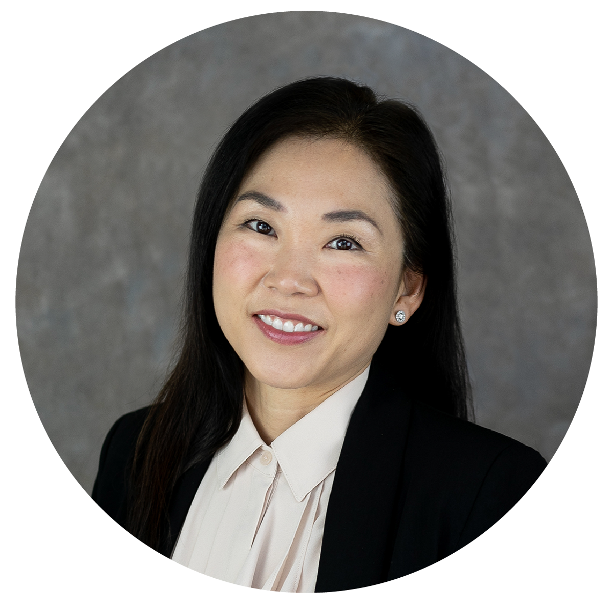 Professional headshot of a woman with dark hair, wearing a black blazer and white blouse, smiling against a gray textured background.