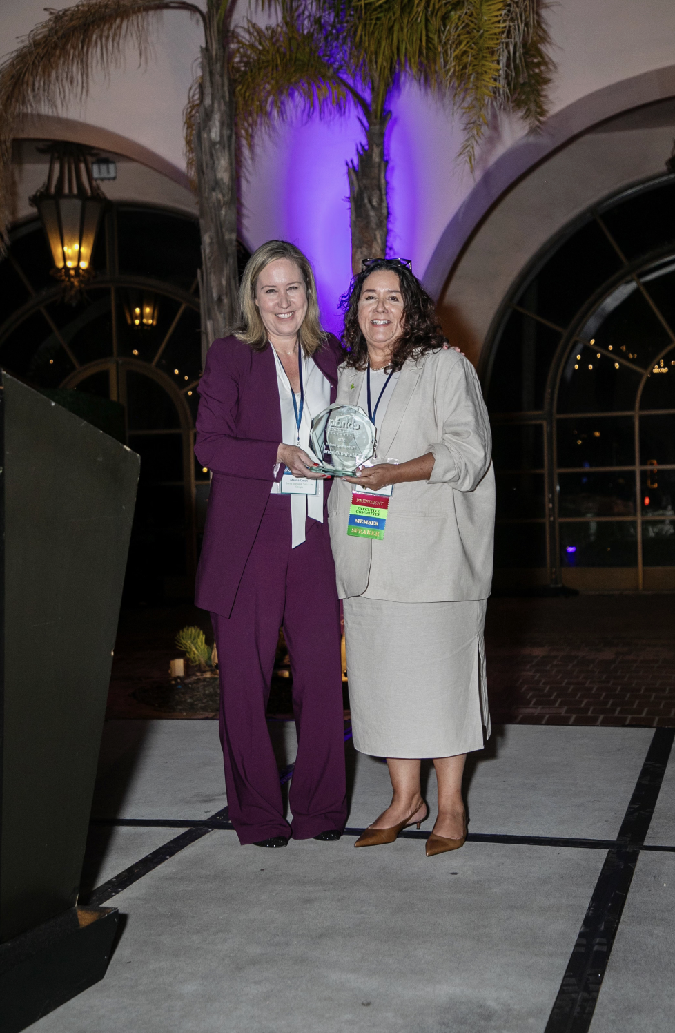 Two women standing indoors holding an award, smiling for the camera. One is wearing a purple suit, and the other is in a beige suit with a skirt. There are palm trees and purple lighting in the background.