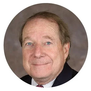 Headshot of an older man with light brown hair, blue eyes, wearing a dark suit and tie, smiling softly against a neutral background.
