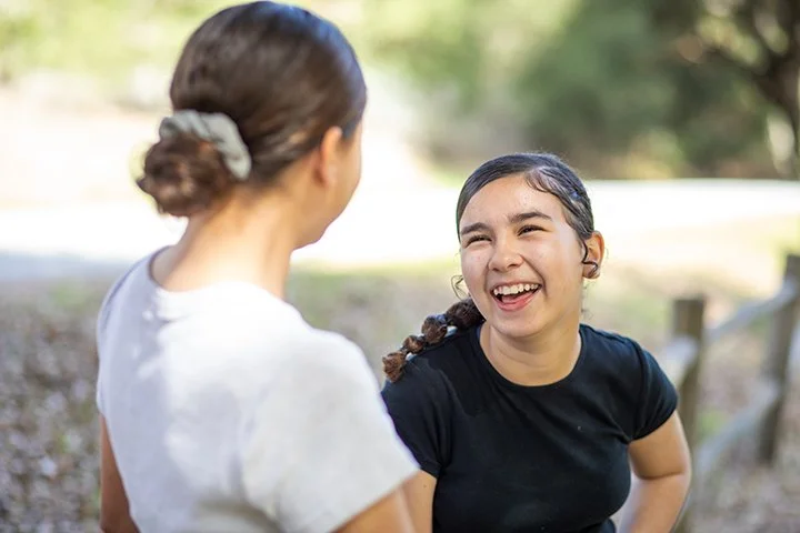 A young girl with dark hair in braids, wearing a black shirt, laughing and talking to an older woman with her hair tied back, outdoors on a sunny day.