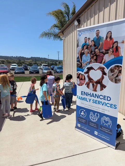Children in line outside a building next to a sign for Enhanced Family Services, with images of diverse families and caring hands, and a parking lot in the background.