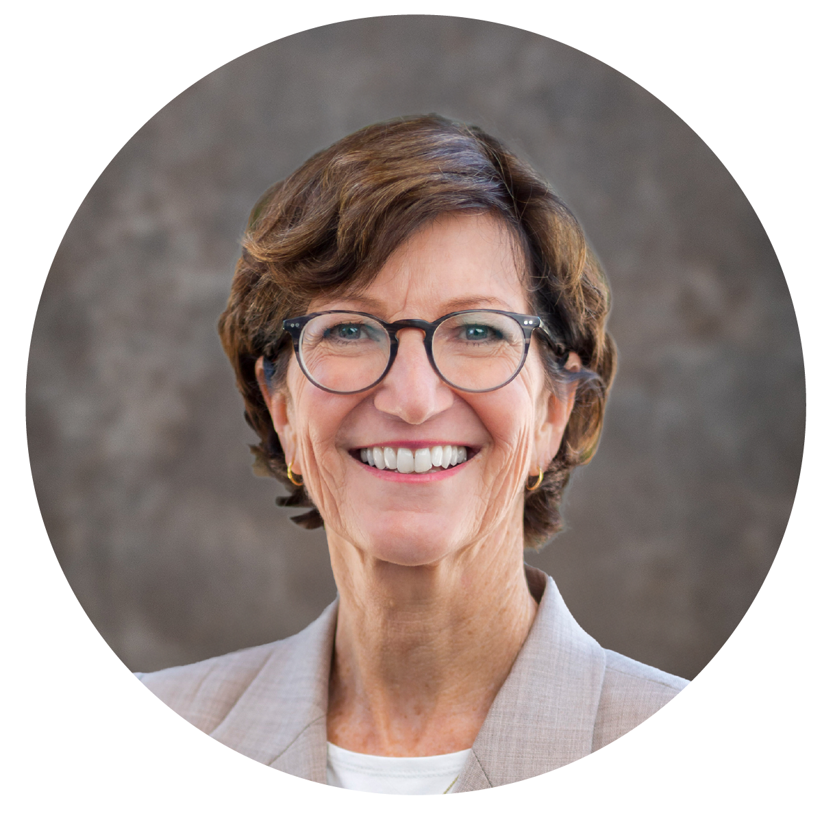 A professional middle-aged woman with short brown hair, glasses, and a bright smile, wearing a beige blazer, in front of a neutral gray background.