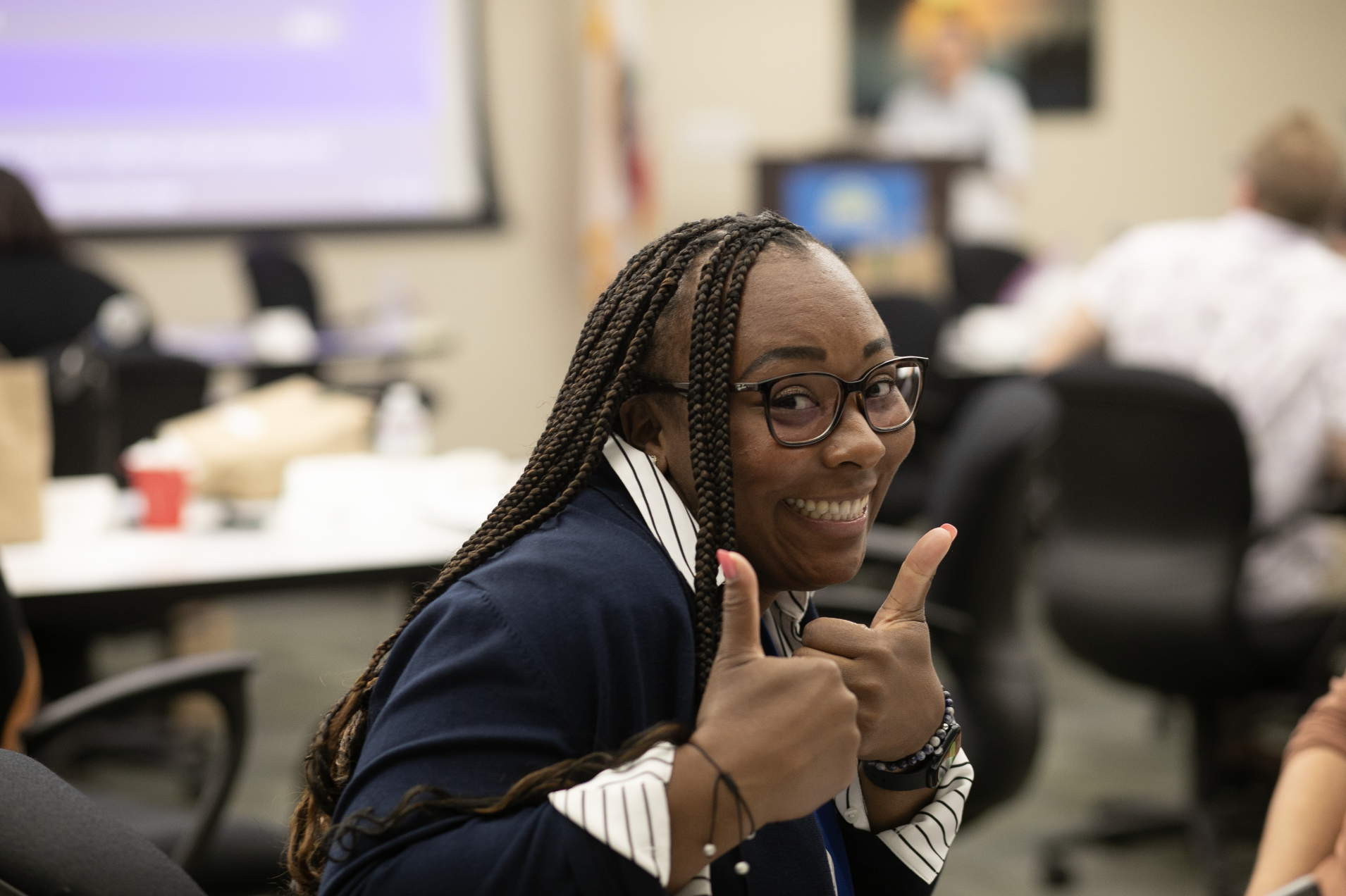 A smiling woman with glasses and braided hair giving a thumbs-up in a conference room.