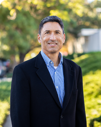 A smiling man in a black blazer and blue checkered shirt standing outdoors with trees and greenery in the background.