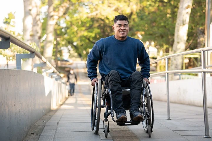 Man in a wheelchair outdoors on a pathway with trees in the background, smiling.