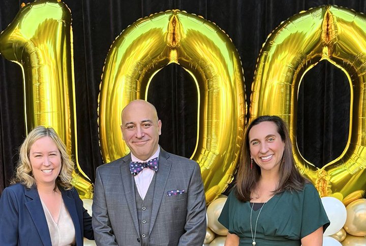 Three people standing in front of large gold balloons shaped as the number 100, celebrating a 100th milestone.