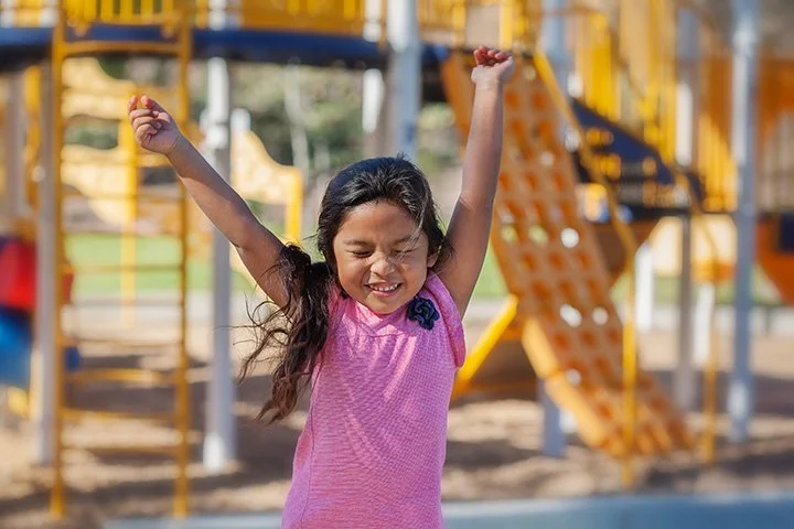 A young girl in a pink dress with a flower brooch smiling and raising her arms in excitement at a playground.