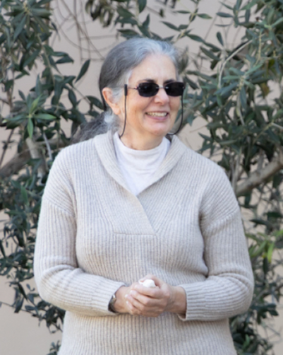 An elderly woman wearing sunglasses and a beige sweater, smiling outdoors with greenery in the background.