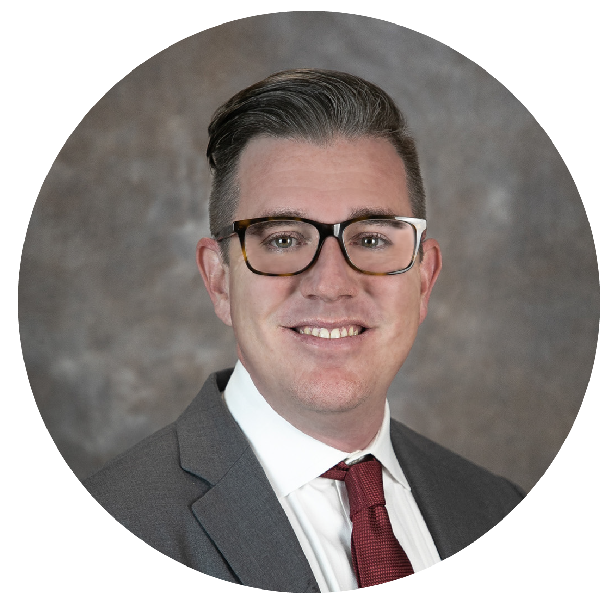 Professional headshot of a man with dark hair styled back, wearing black glasses, a dark suit, white shirt, and maroon tie, smiling, against a neutral background.