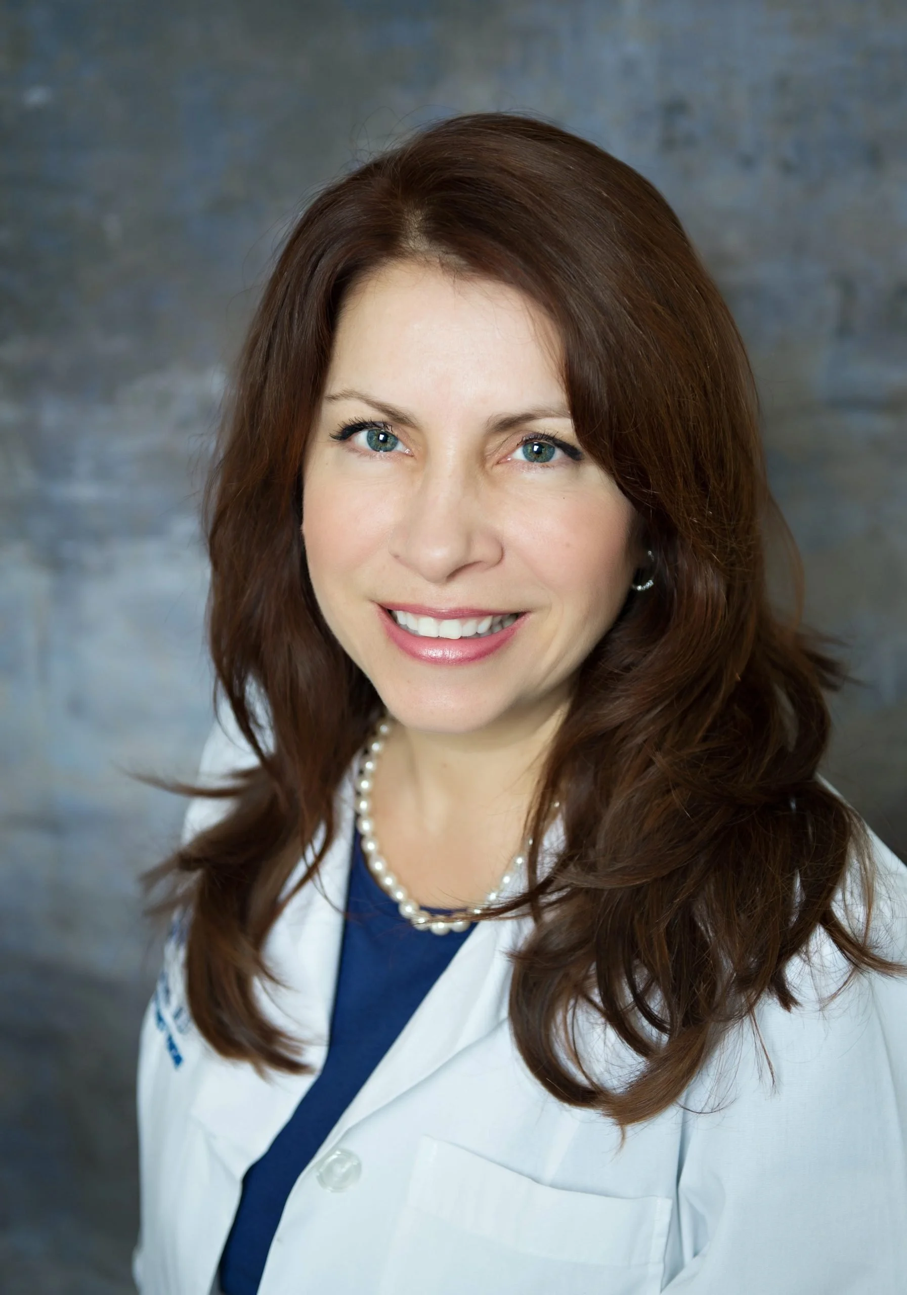A woman with long, wavy brown hair, blue eyes, and a warm smile. She is wearing a white lab coat, a blue top, and a string of pearls. The background is a textured gray surface.